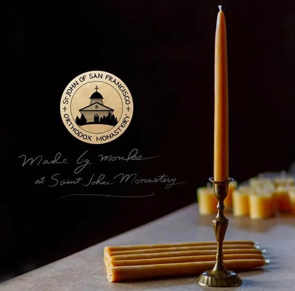 A set of tall, thin beige dinner candles placed on a surface with a candle holder, with the logo and text indicating they are made by the monks of St. John's Monastery.