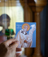 Hand holding a card with a religious illustration of a saint against a blurred background