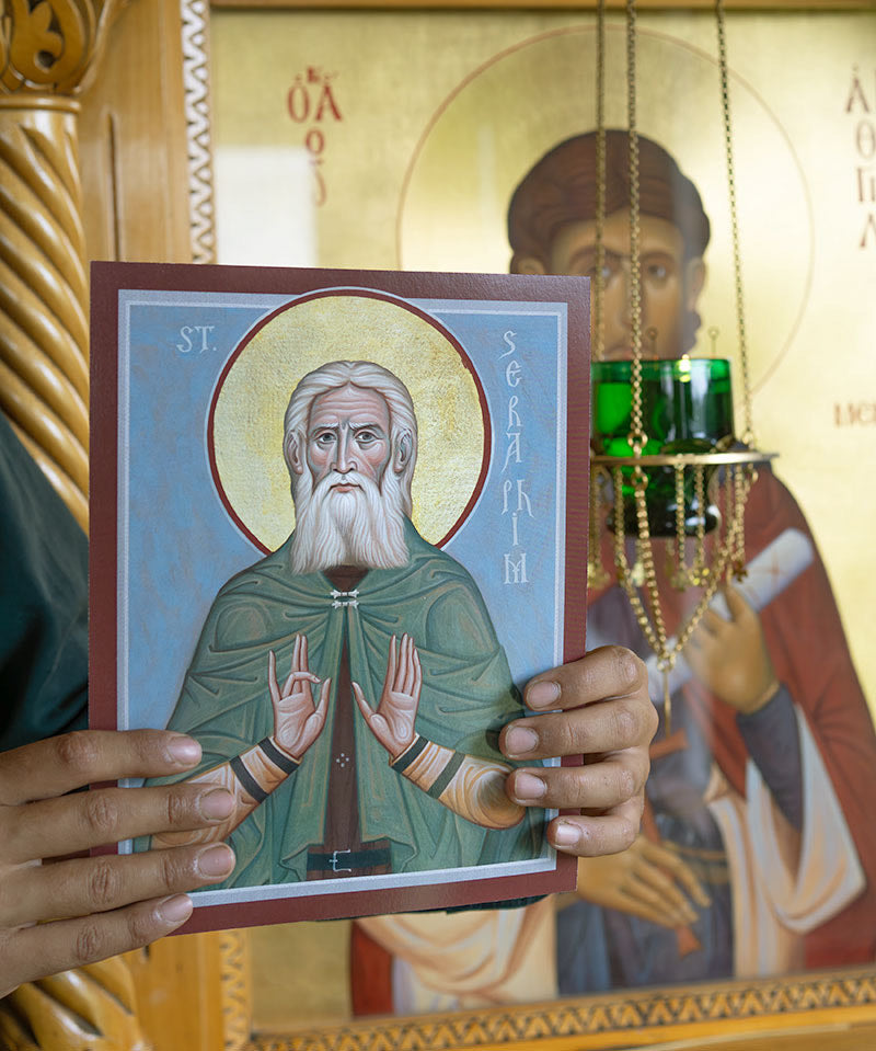 Person holding a icon of St. Seraphim of Sarov in front of a larger religious painting, which is a an icon of St. Agathangelos of Esphigmenou